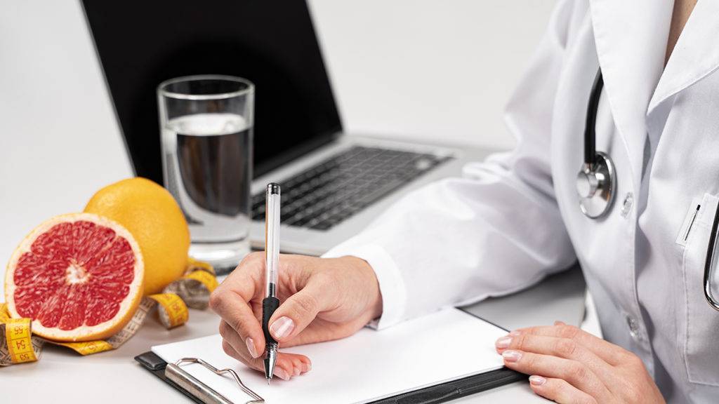 Nutricionista faz anotações em um papel, no fundo há uma grape fruit, um copo de água e um notebook.